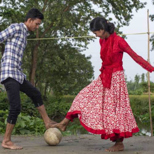 A boy and a girl play woth a football on a dirt path with greenery behind them.