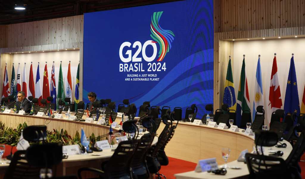 A large banner of the G20 Brazil 2024 Summit logo with country flags either side, in a large meeting room with a row of chairs in front of a long, curved desk.