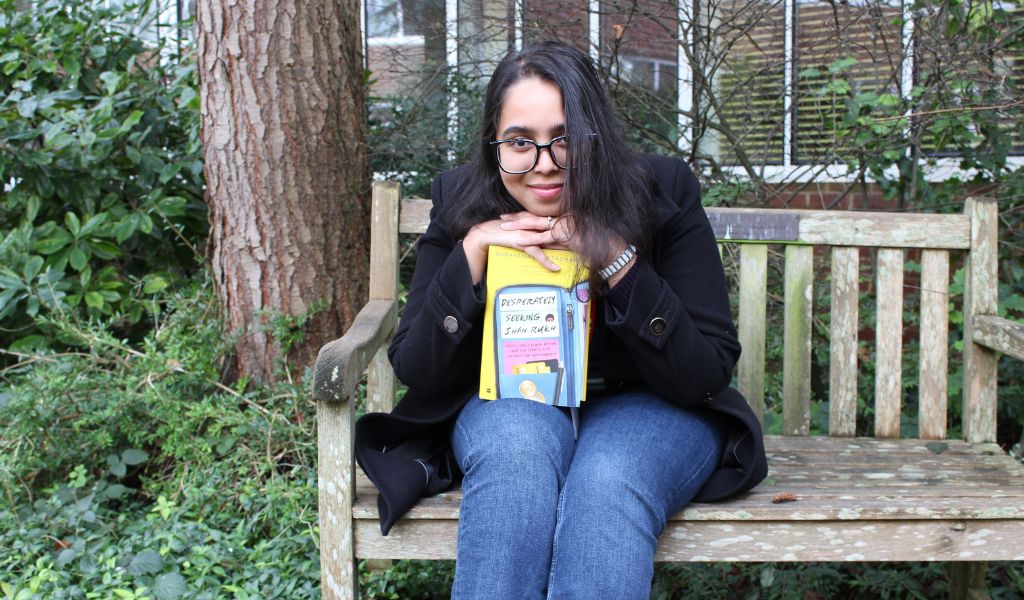 A female with long dark hair and classes, wearing a black jacket and blue jeans, sits on a wooden bench, holding a book.