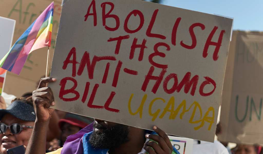 ‘Abolish the anti-homo bill’ is written in red and ‘Uganda’ written in yellow on a cardboard sign, held by a black man wearing a white t.shirt and a rainbow coloured scarf. Behind him a re more people holding cardboard signs. Above them is a bright blue sky.