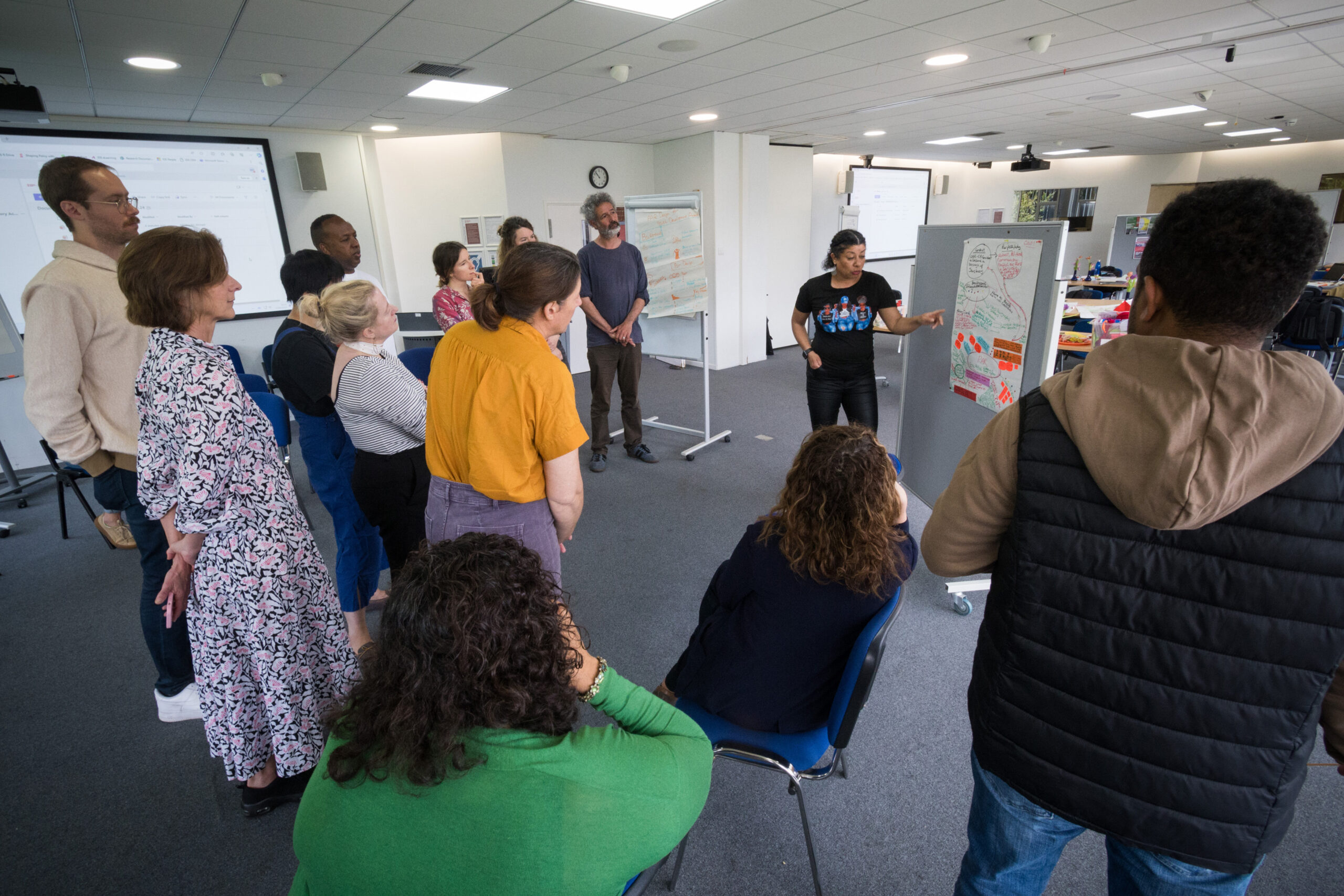 A woman in a black top and trousers stands next to a pin board pointing to a flip chart with written text on it. Several people stand in a circle listening to her speak and looking at the flip chart.