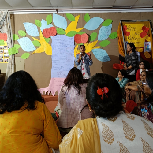 A girl standing on a stage speaking to a group of women. Behind her is a tree made out of pieces of colourful paper with text on it.