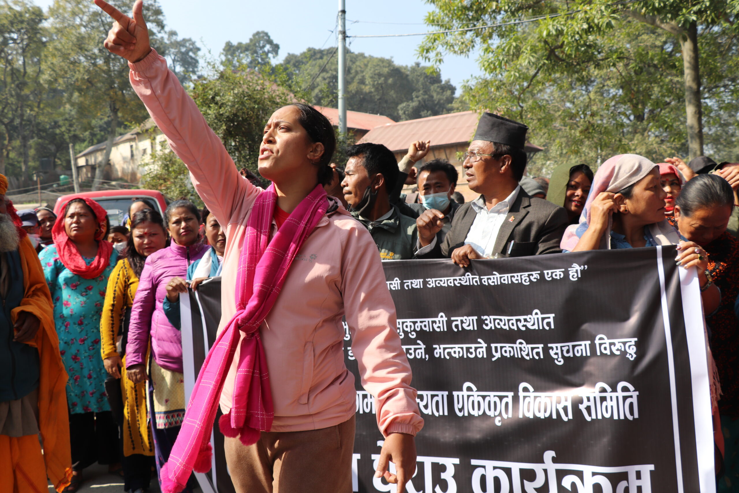 A woman in a pink top and scarf stands at the front of a protest with her arm raised and her hand pointing up. Behind her, several people are carrying a black banner with white text.