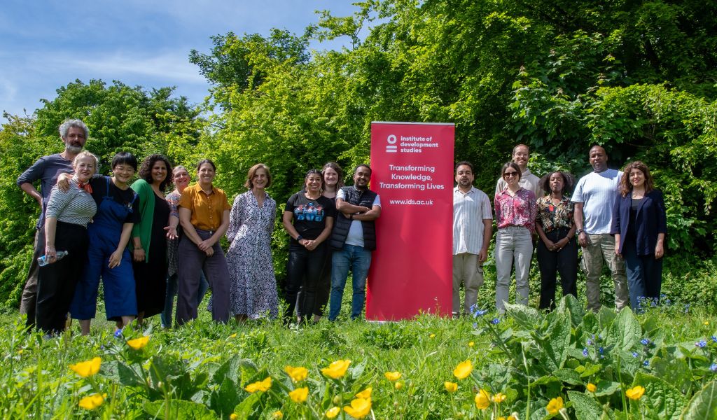 A group of people standing in a line facing the camera on grass. A red roller banner is in the centre, which carries the text "Transforming Knowledge, Transforming Lives."