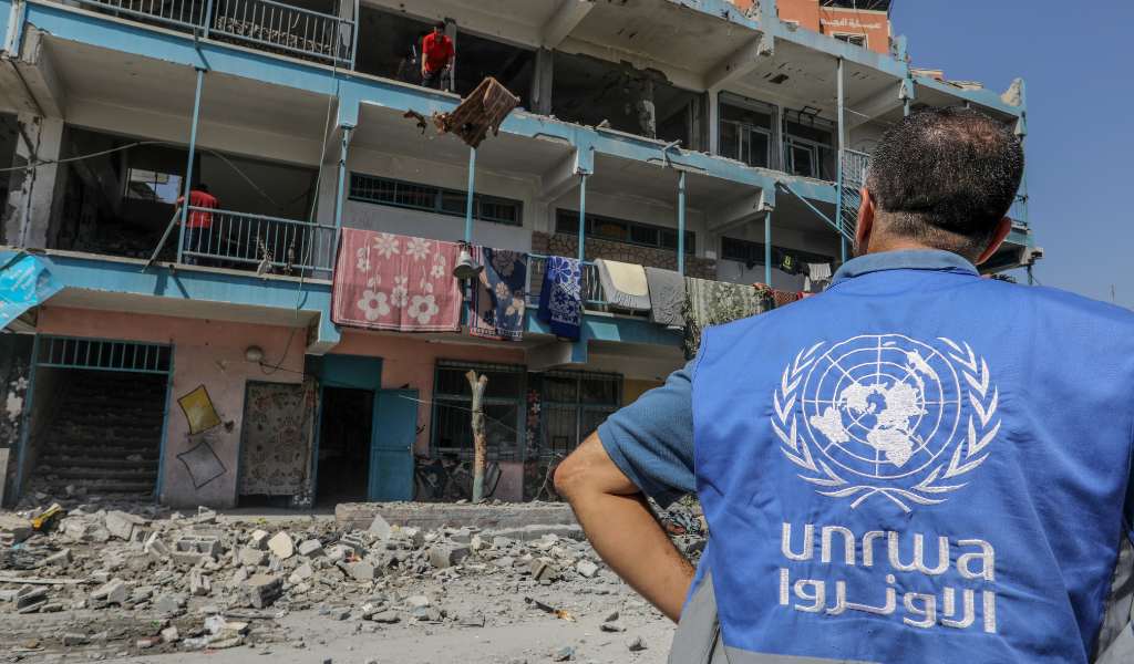 The back of a man's blue shirt showing 'UNRWA' and its logo. He is looking at a three storey building with all the windows blown out and rubble at its base.