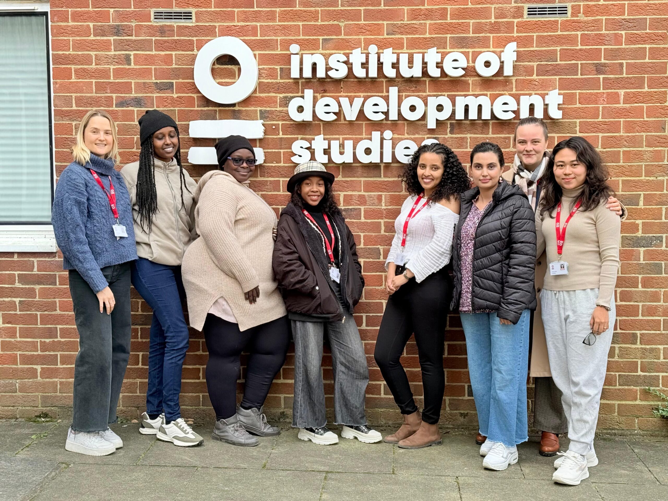 8 women stand together outside a brick building with a sign in white writing saying Institute of Development Studies. The women wear clothing like jeans and light jackets.