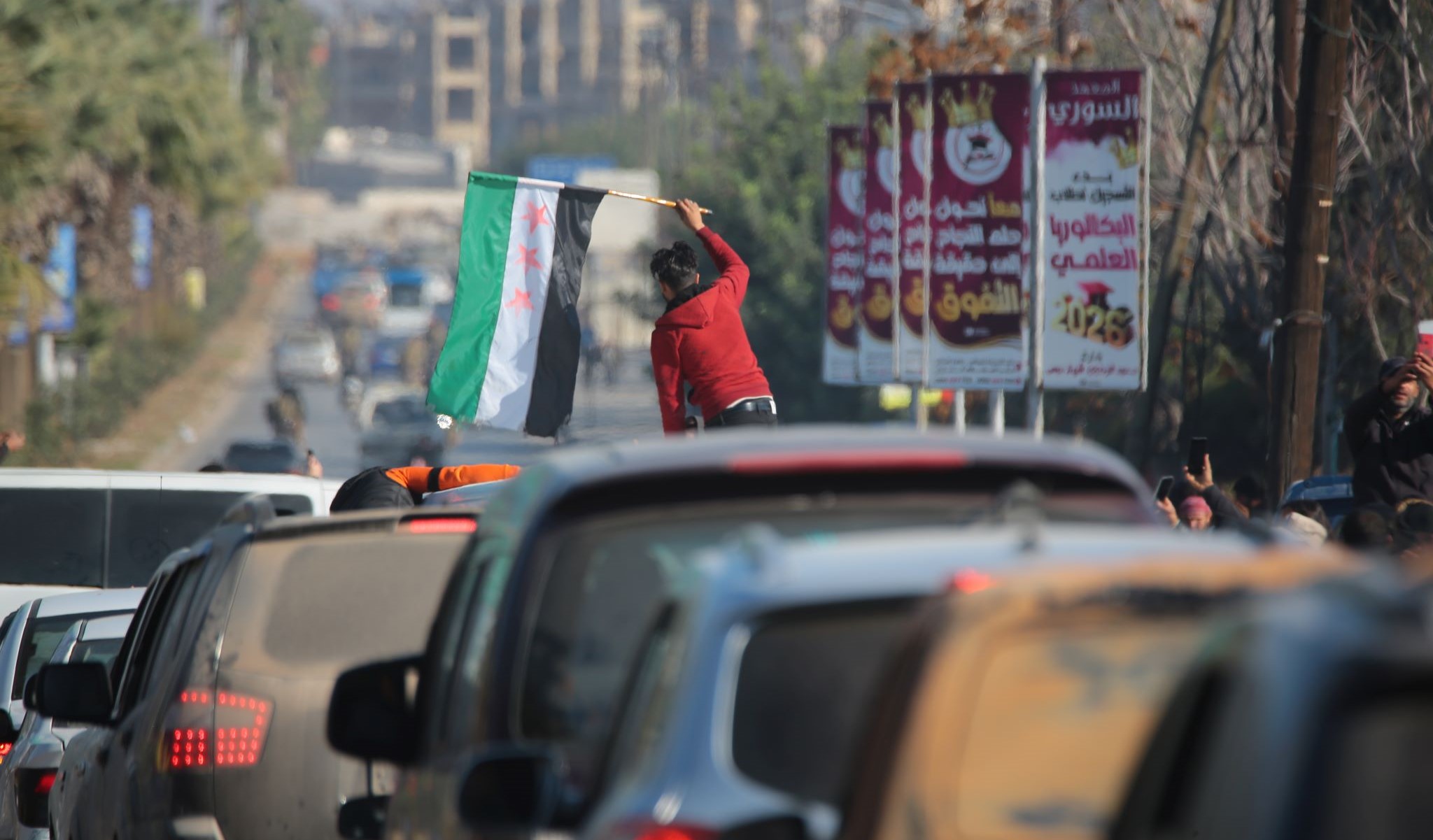 A line of cars, with someone standing up in a car waving the new Syria flag