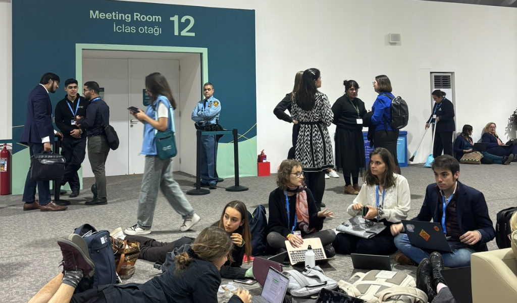 Young people sitting with badges outside a room with entrance coloured blue and titled Meeting Room 12.
