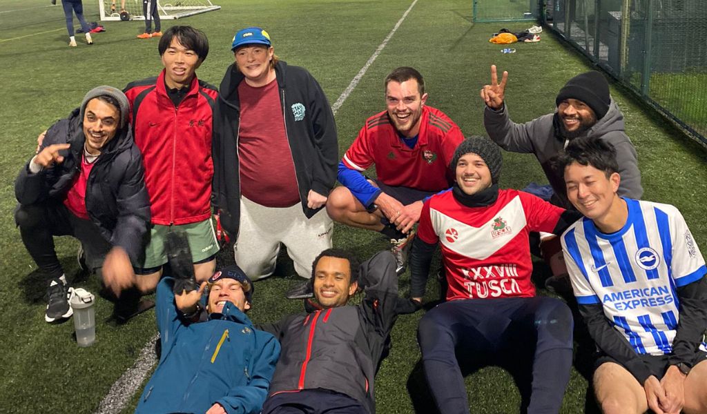 Nine students all in sportwear, on a football pitch. The two in front are laying down. They are an assortment of different nationalities and genders.