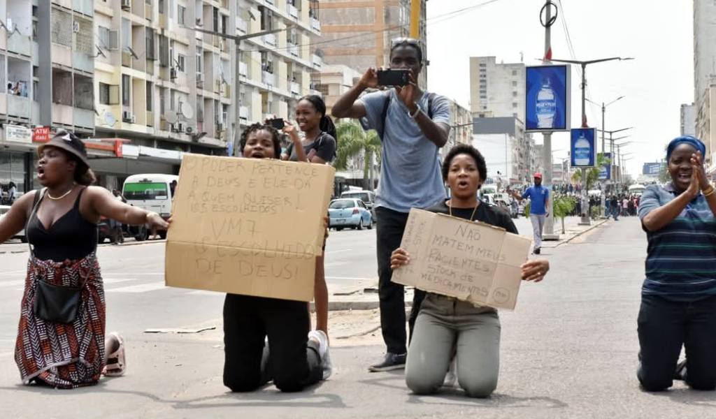 Four women in a row on their needs on a concrete pavement. Two of them in the centre hold hand written cardboard signs.