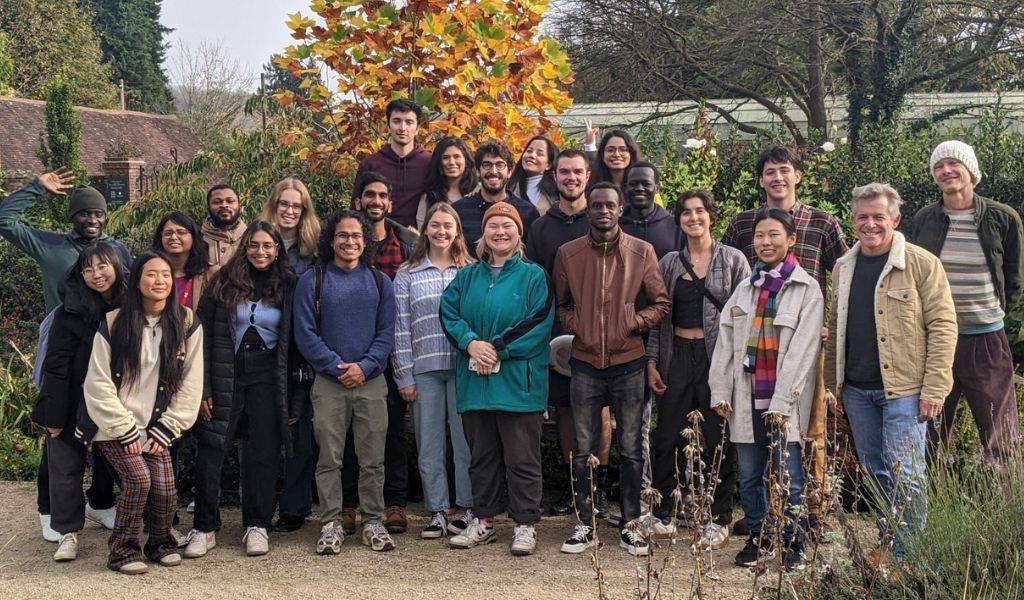 A group of 24 people smiling at the camera, standing in front of a tree with orange leaves.