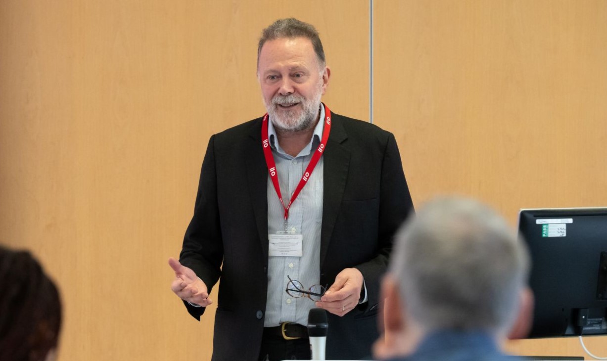 A man wearing a jacket standing and appearing to be talking to an audience.
