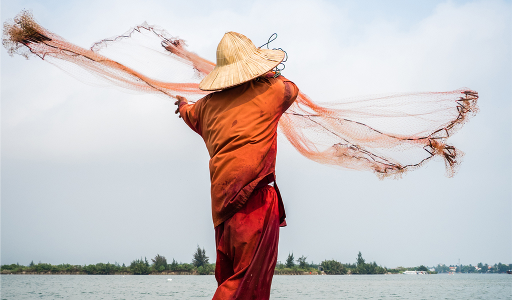 A fisherman with his back to us wearing an orange garment and a straw hat throws a fishing net into the sea.