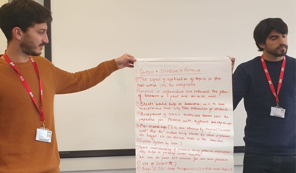Two young men standing in a classroom, holding up a sheet of flip chart paper with writing on it.