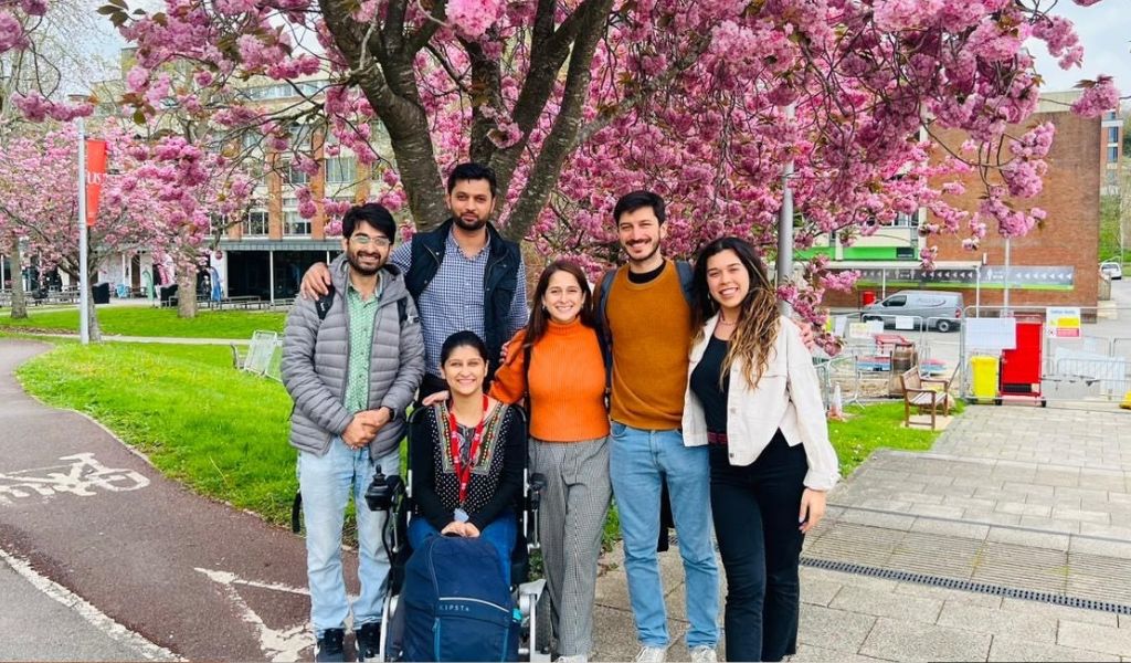 A group of six students, a mixture of males and females, standing in front of a tree with pink blossoms.