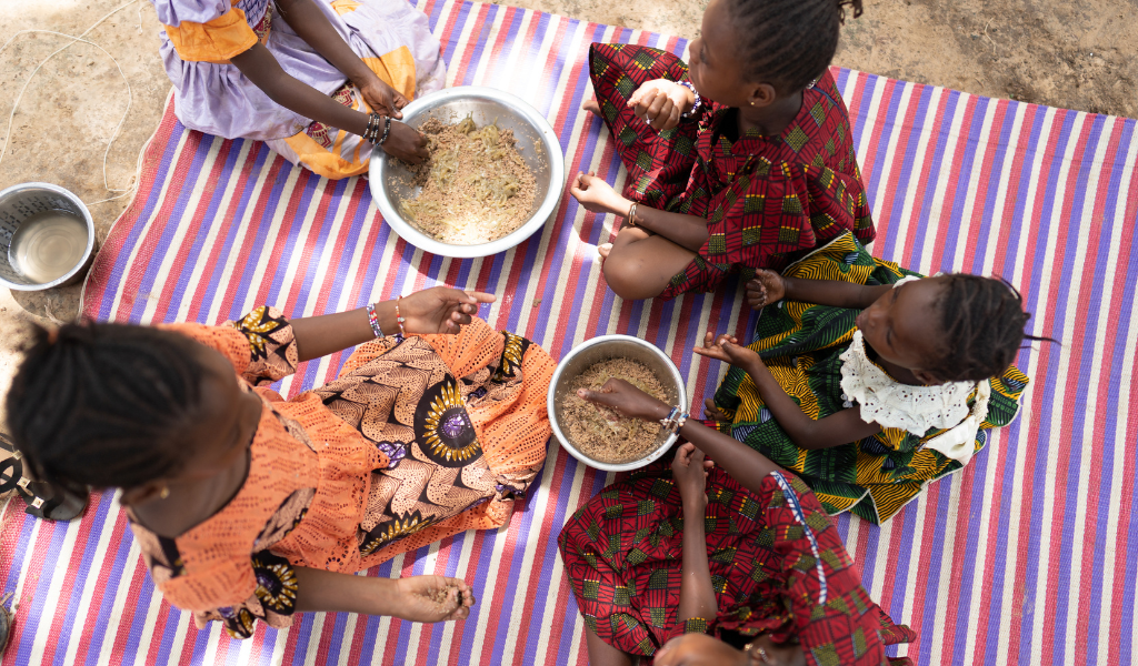 Girls wearing colourful dress sit on a red and white stripe map with brown colour cereal in bowls.