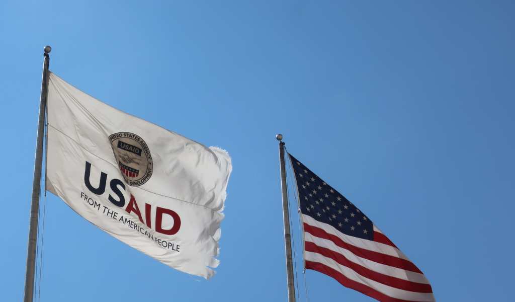 Two flags flying on flagpoles against a blue sky. The left flag is the USAID logo. The one on the right is the USA country flag.