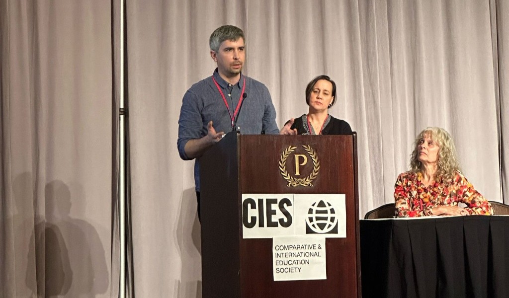 A man stands behind a lectern on a stage, giving a speech. A woman stands beside him, with another woman sitting down on a table. The acronym CIES is visible on the lectern.