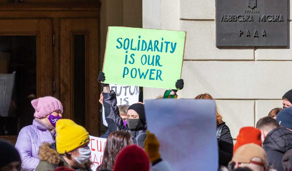 A woman in a crowd holds up a banner saying 'Solidarity is our power'. She wears a mask covering her face and a black headscarf.