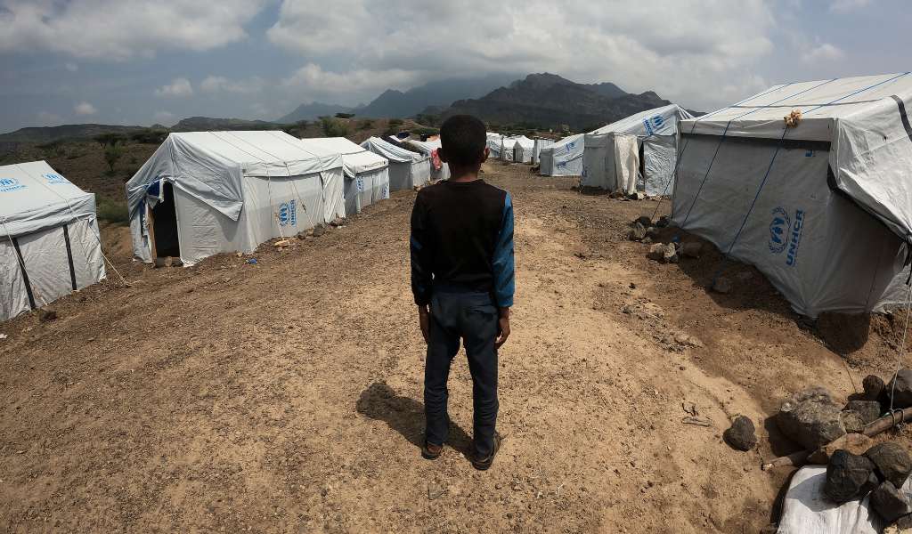 A boy dressed in dark clothing stands with his back to the camera looking at a row of white tents. The ground is like sand and the tents have the UNHCR logo stamped on them.