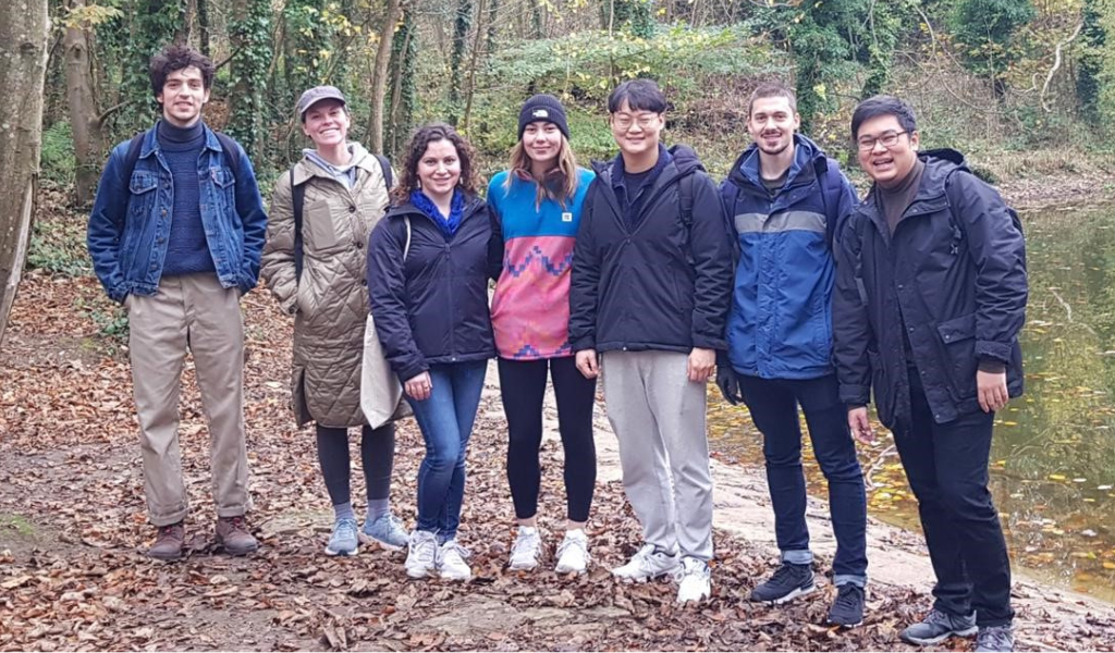A photograph of a group of seven students standing in a wooded area. They are different genders and nationalities.