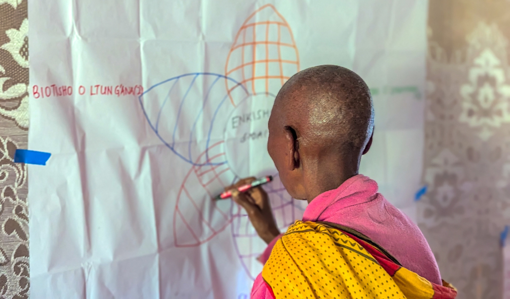 A woman with her back to us uses a marker pen to write on a flip chart on a wall. She is wearing bright yellow and pink.