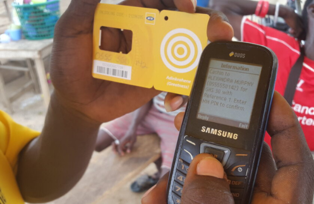 A hand holding a basic Samsung phone showing a text confirmation of mobile money load.