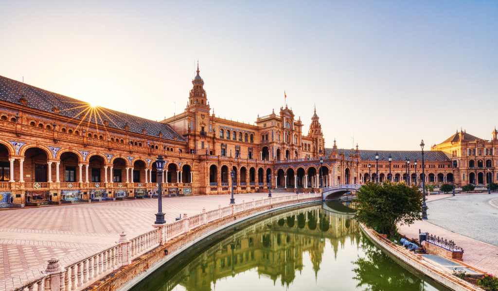 A grand, sweeping, historical building with many arches, with the sun setting behind it and a strip of wide pavement and canal running in front of it.