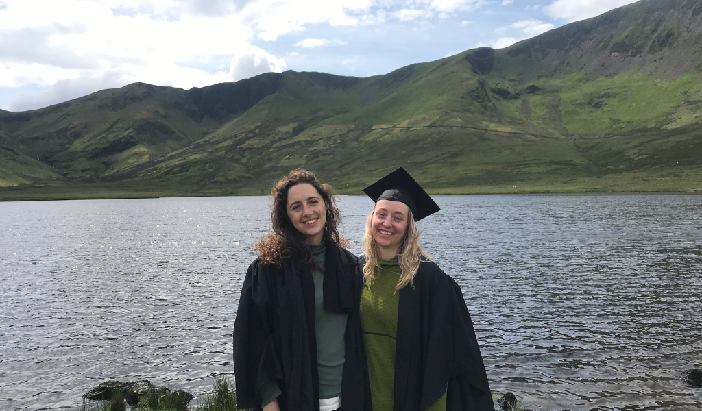 Two women wearing black graduation caps and gowns stand next to each other smiling. Behind them is a lake and some green mountains.