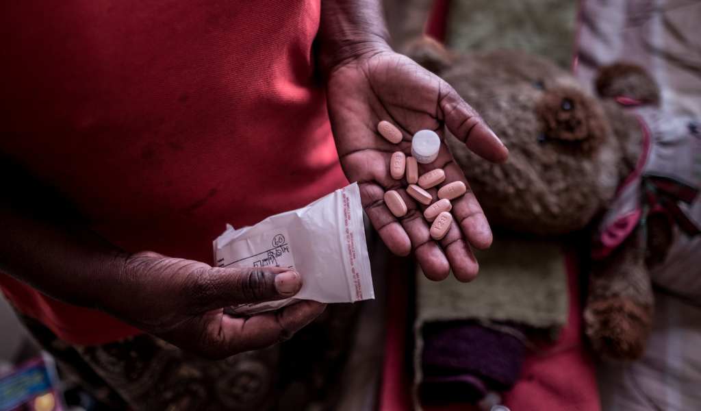 A hand held out with a selection of pills in the palm. The person wears a red t.shirt and the packet the pills have come from are in the other hand.