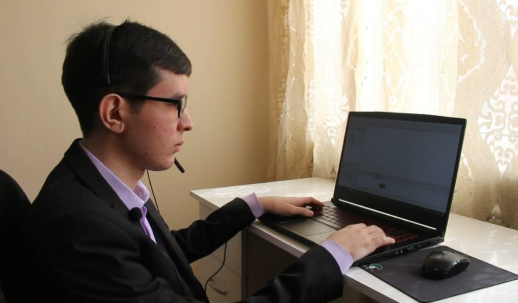 A photograph of a man wearing a purple shirt and a black jacket, sitting at a laptop with his hands on the keyboard. He has glasses on and is also wearing a headset.