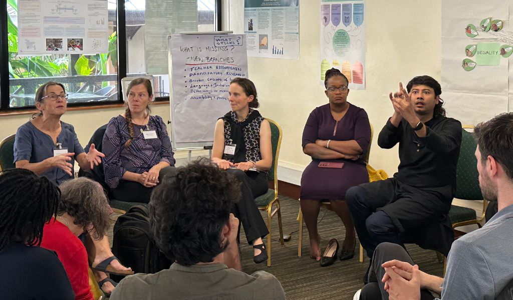 A photograph of researchers sat on chairs in a circle during a workshop. The focus is on five people sat next to each other. The left-most person is speaking and gesturing with their hands, and the two people to their left are concentrating on them. The right-most person is delivering Sign Language Interpretation for the discussion.