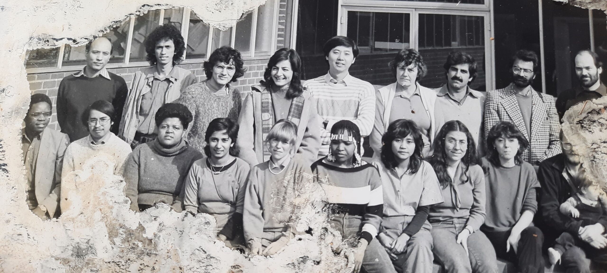 A black and white photo of a group of 20 people. The photo is old and has some damage to the edges.