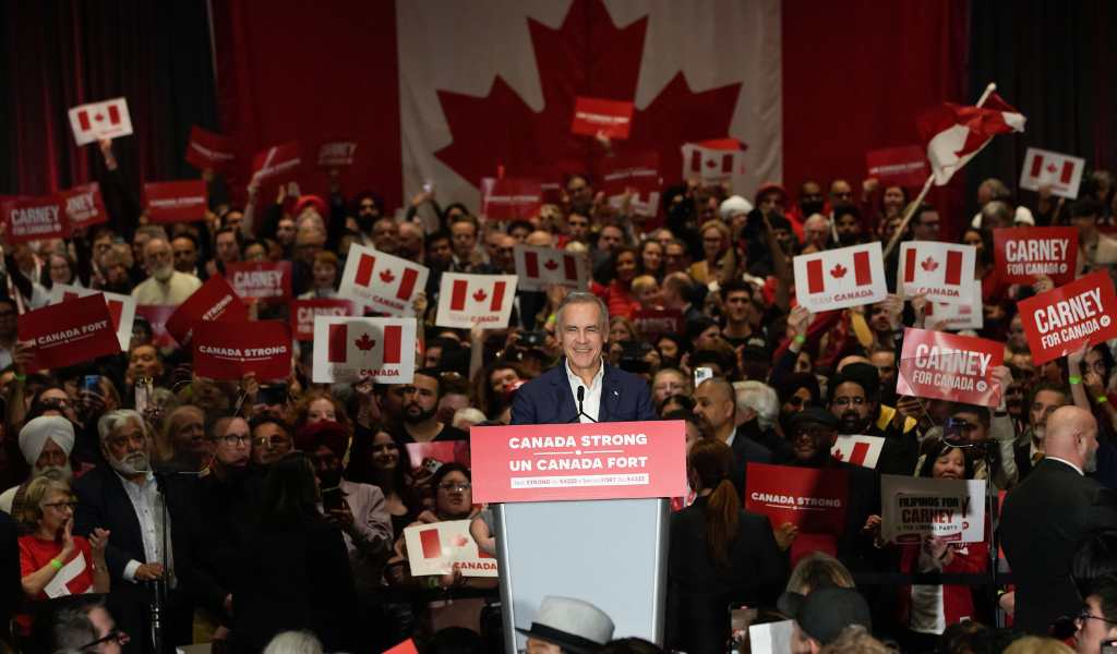 Mark Carney stands at a podium in front of a large crowd holding up images of the Canadian flag, in a dark hall, with the maple leaf flag hanging in the background.