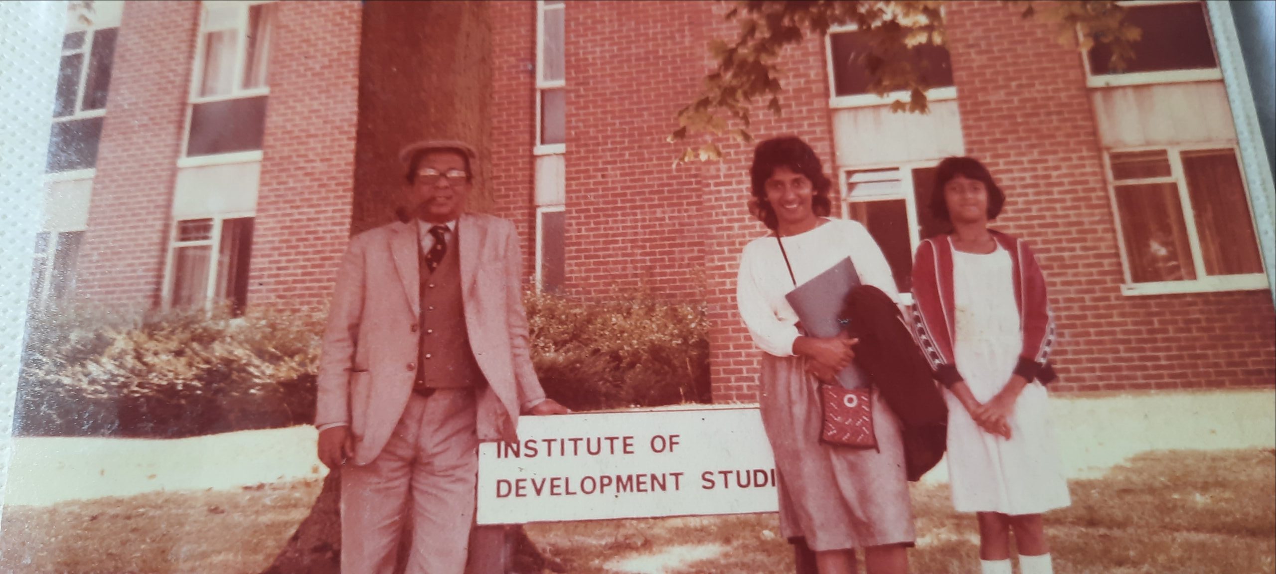 An old colour photo of a man and two young women standing on either side of a sign that says 'Institute of Development Studies' in front of a brick building.