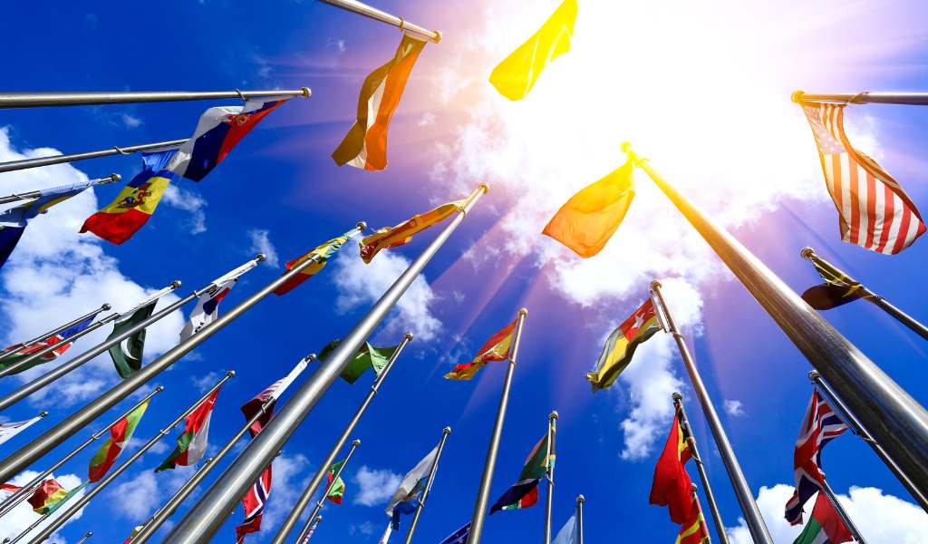 Multiple flag poles with various country flags photographed from below. Above them is a blue sky with a blurred sunlight bursting through in the top right corner.