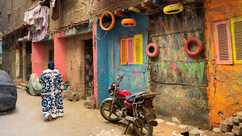 A woman in a blue and white patterned long dress walks past colourful houses in Cairo, Egypt. A motorcycle is in the foreground.