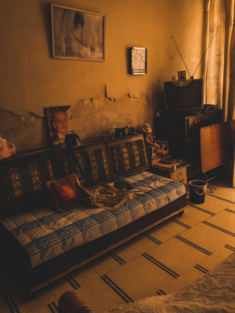 Damascus image A photo of a sofa covered in white and blue fabric, with a photo of a man propped up behind it and television on a stand on the right-hand side. The plaster on the wall behind it looks like it has fallen off.