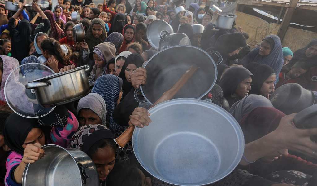A crowd of people squashed together holding up large metal bowls, desperate for food aid. Many are wearing headscarves in dark blue, grey and black colours.