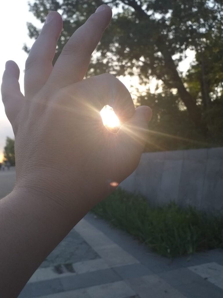 A close-up photo of someone’s left hand, with the finger and thumb making a circle, and the sun shining through the gap. There is a tree in the background.