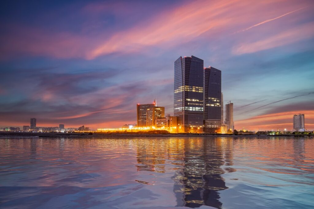A skyline at sunset, with two tall buildings in the centre and a row of bright lights at the bottom. A stretch of water is in the foreground.