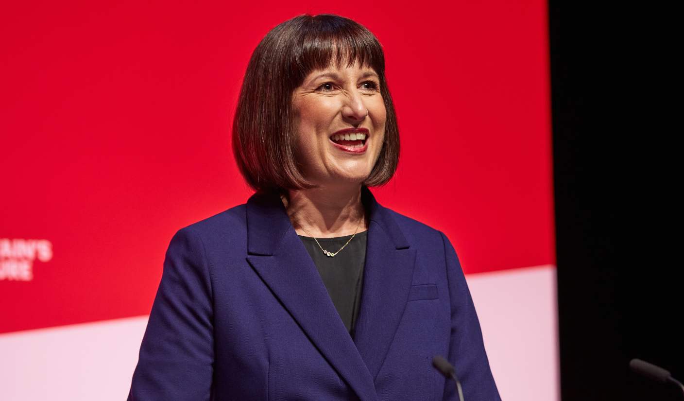 Rachel Reeves from the waist up wearing a navy blue jacket, smiling, with a red screen behind her.