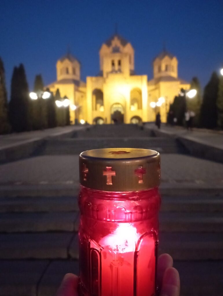 A photo of a grand-looking church is in the background, blurred. In the foreground is a hand holding a red glass jar with a candle in it, which is in sharp focus.