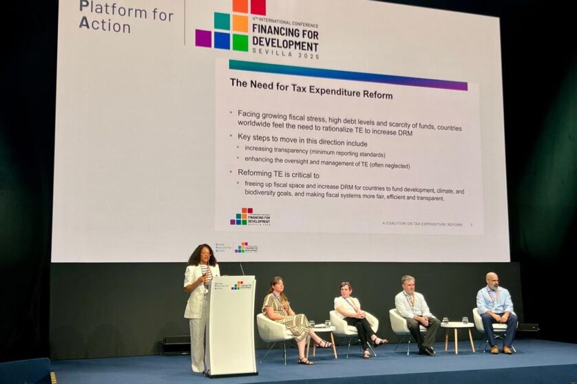 A woman stands at a lectern with a text-based slide behind her. Four other people sit behind her on the stage