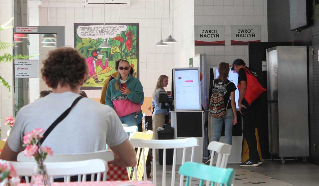 A busy restaurant scene, one person in a white t.shirt sitting at a table with their back to the camera. Other people standing about to exit, others standing at screens to order food.