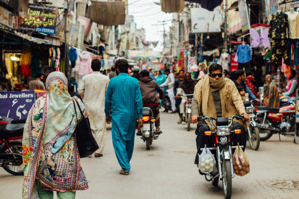 View of a busy market street in Pakistan with people walking in the middle and a man in on a motorbike facing the camera. To the left and right, market stalls with clothes on display. 