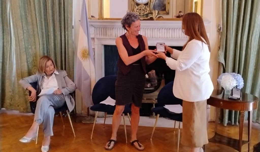 A woman dressed in black receives an award from a woman wearing a white jacket. Both are standing inside, in front of a large fireplace
