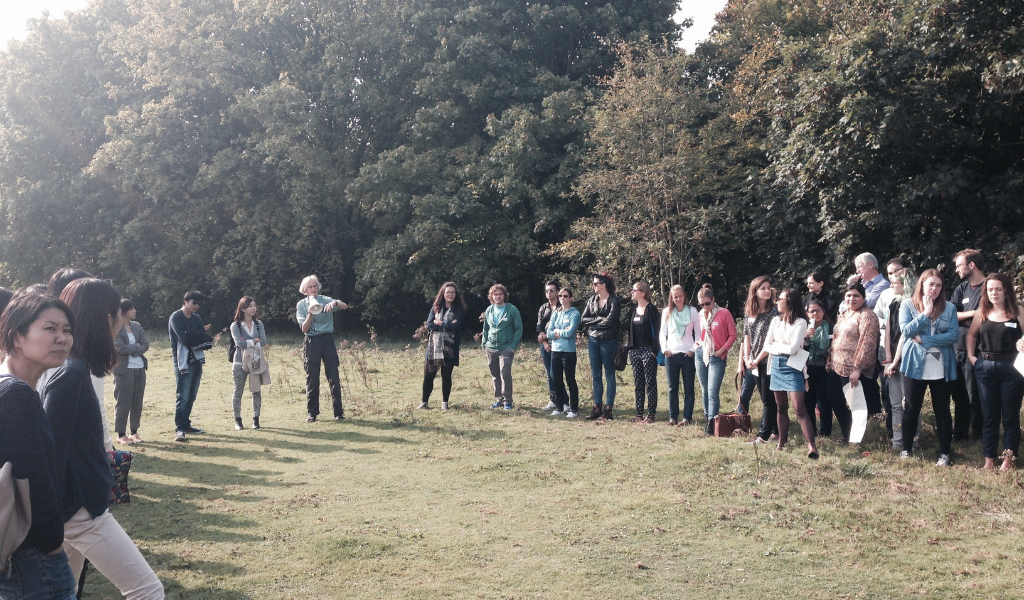Approximately 20 people standing on the grass in a semi-circle. One person is speaking and the rest are listening. There are trees behind them and the sun is shining.