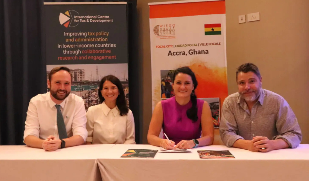 Four people sitting in a row behind a desk, smiling at the camera while signing a document.