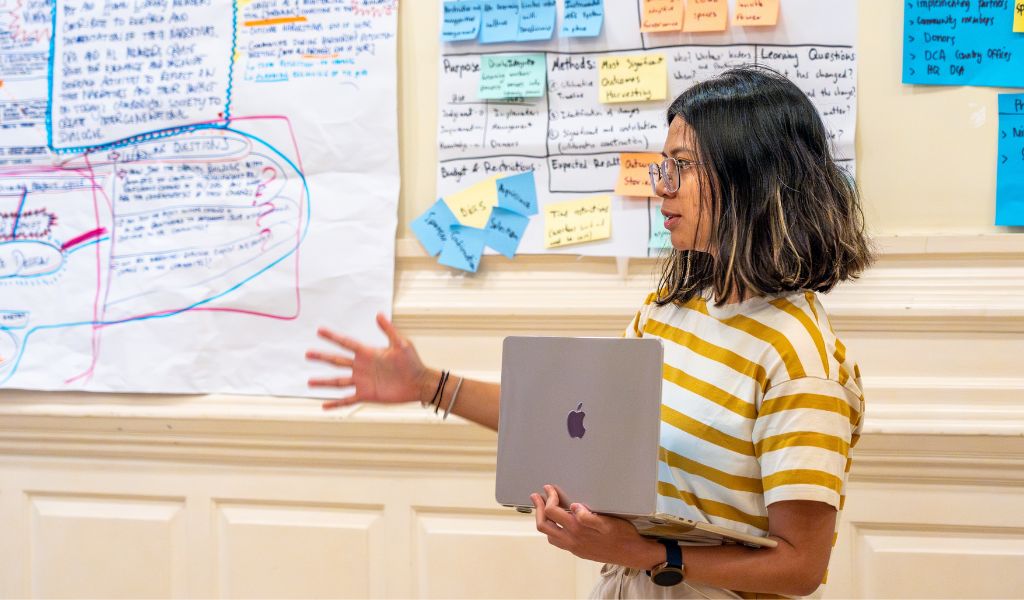 A photo of a person with shoulder length hair and a stripy yellow tshirt, holding a laptop. A number of postit notes with writing on them are stuck up in the background.